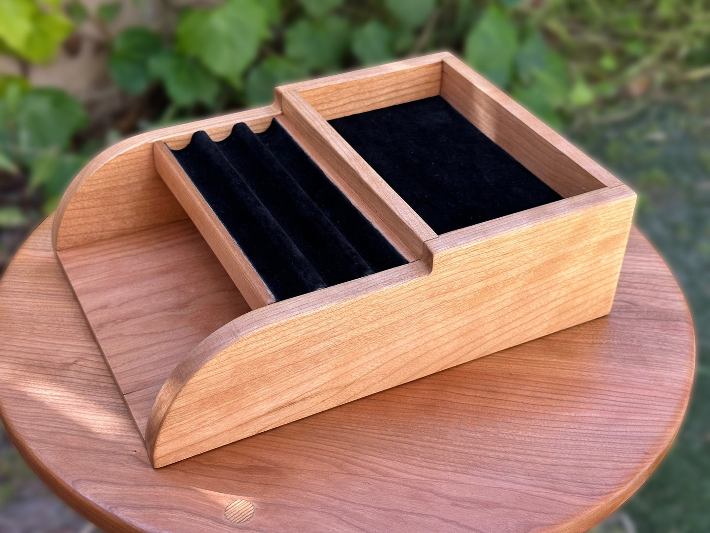 A handcrafted cherry wood desk organizer with curved side panels, black velvet-lined slots for pens, a spacious rear compartment for ink bottles or accessories, and a lower flat section for holding notes or papers, shown on a round wooden table outdoors with green foliage in the background.