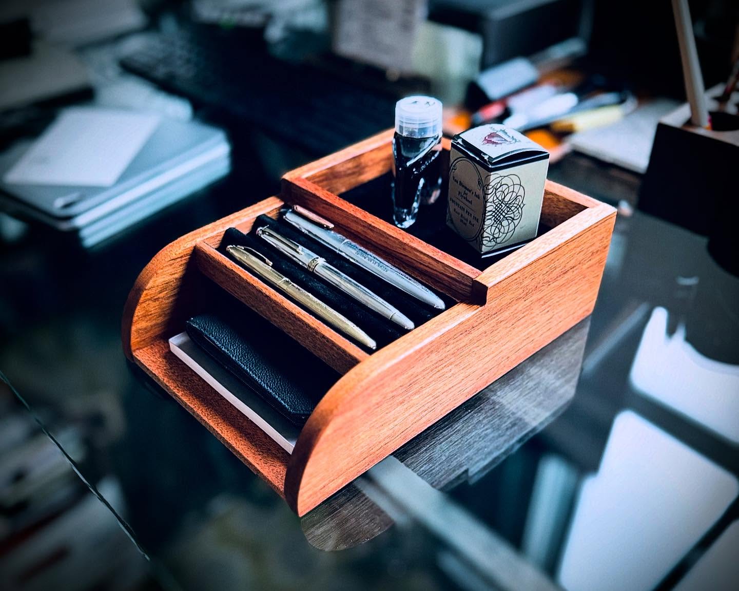Mahogany desk organizer, Pen Tomo,  holding several modern fountain pens, an ink bottle, and a boxed ink cartridge, with notebooks stored in the bottom slot. The organizer sits on a glass-top desk in a workspace with blurred office supplies in the background