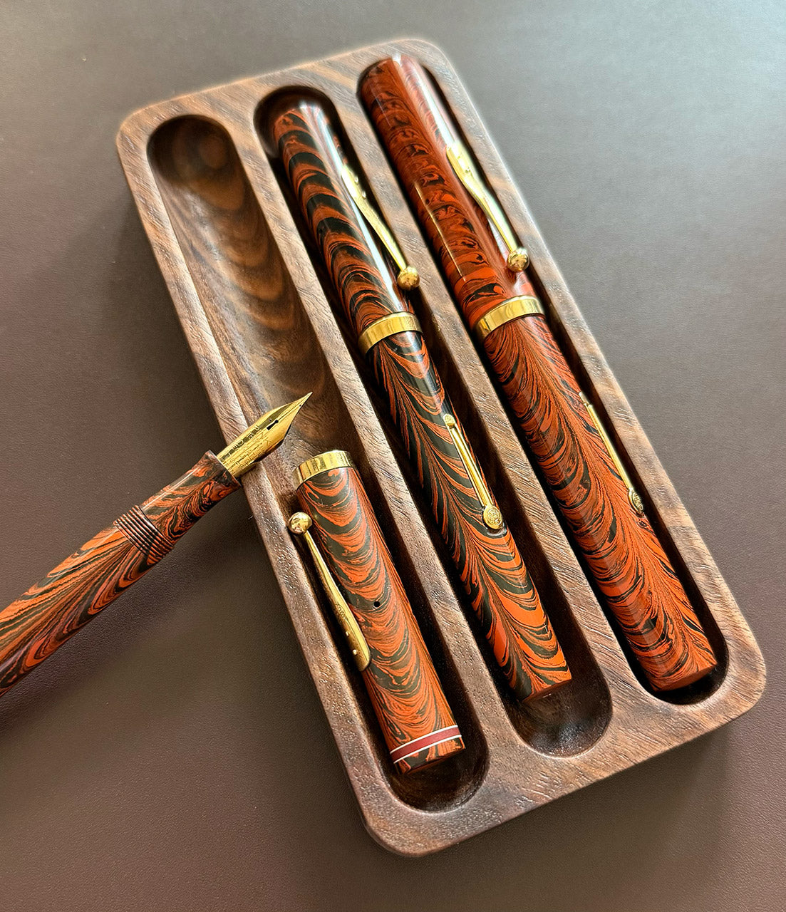 Close-up view of three vintage-style fountain pens with red and black ebonite swirl patterns and gold trim, displayed in a handcrafted figured walnut pen tray