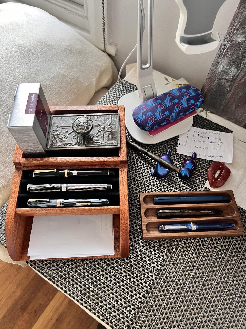 A neatly arranged desktop featuring a wooden pen organizer with three luxury fountain pens and a stack of white paper at the bottom. To the right, a smaller walnut pen tray holds three additional pens. A decorative metal box and a bottle of Yamabudo ink are placed atop the pen organizer.