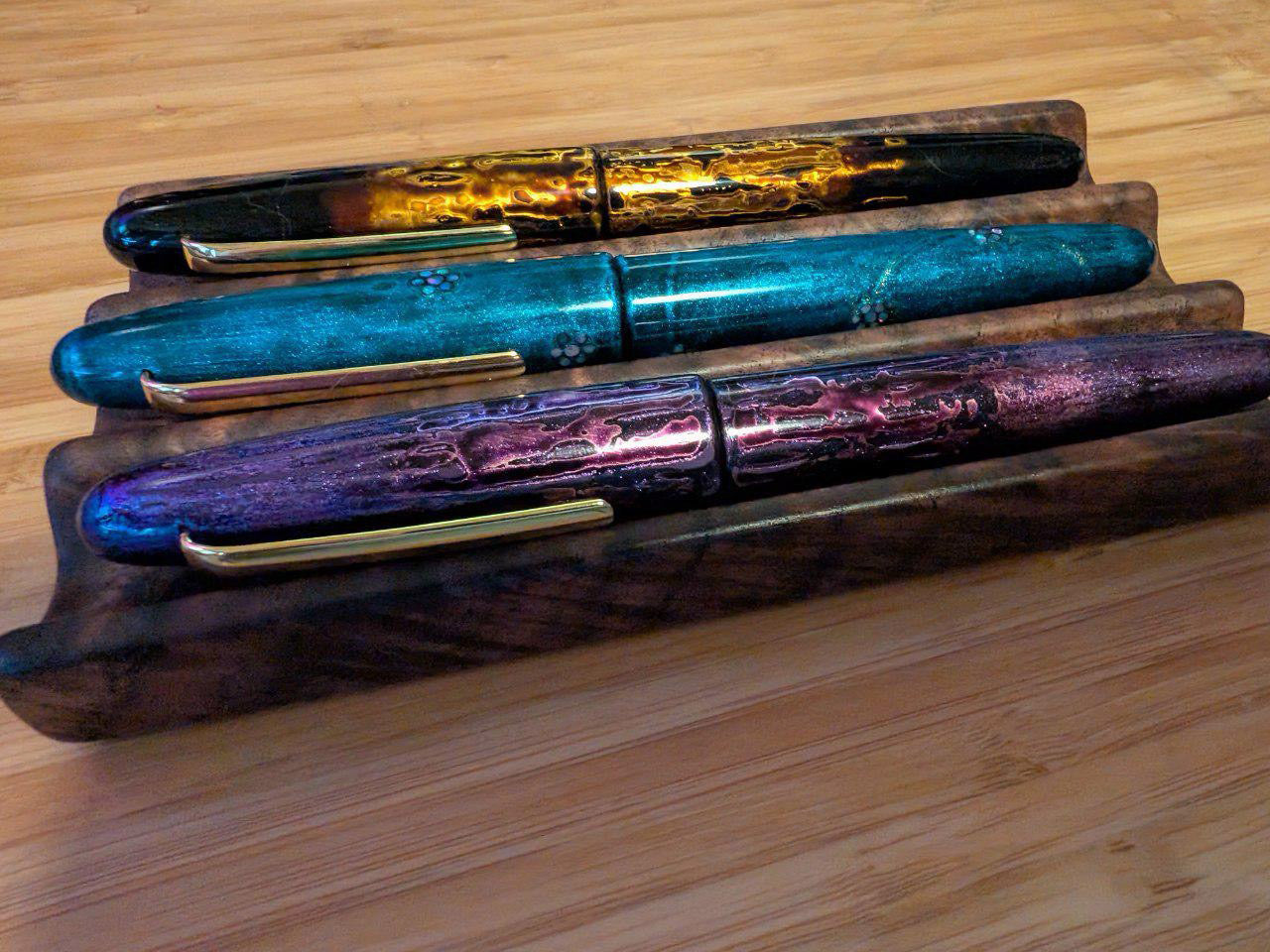 Three iridescent fountain pens with gold clips in shades of bronze, teal, and violet, displayed in a figured walnut pen tray on a bamboo surface