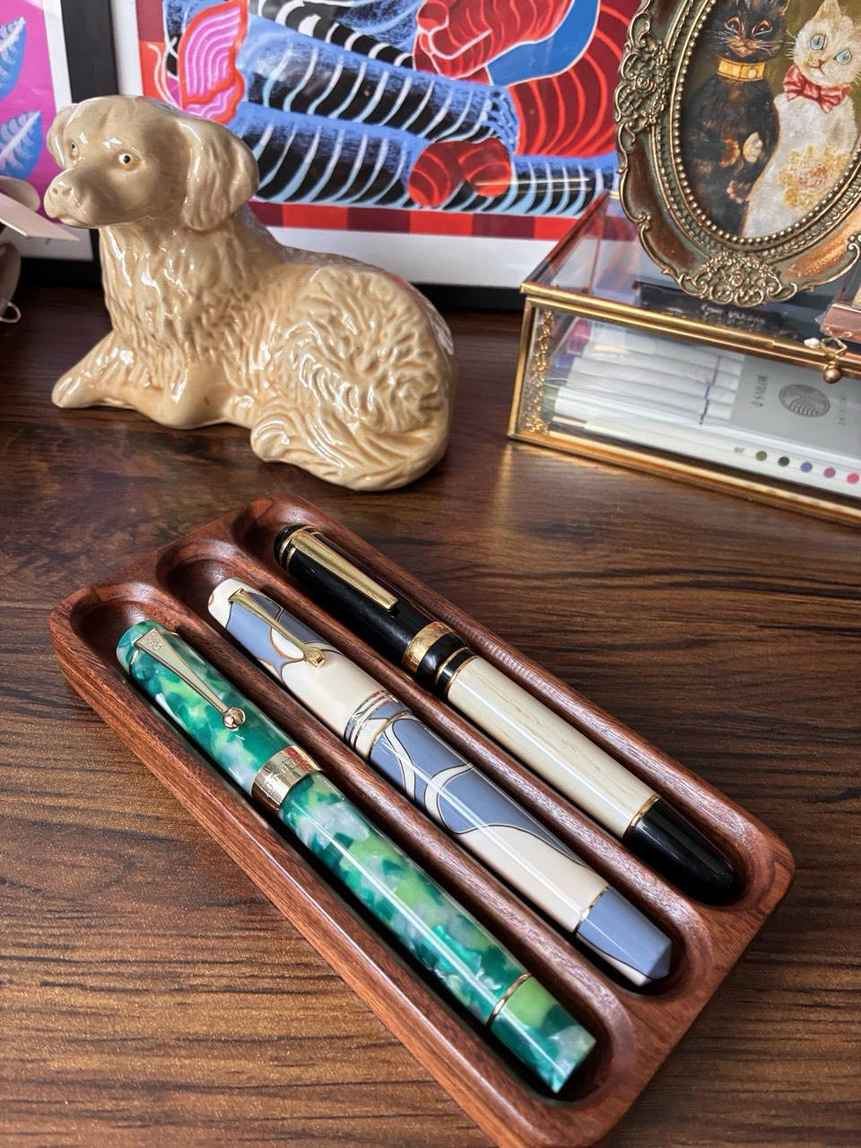 Three elegant fountain pens with marbled and enamel finishes displayed in a walnut tray on a dark wood desk, surrounded by a ceramic dog figurine, framed cat artwork, and colorful art prints in the background