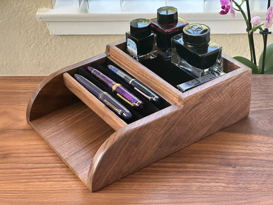 A handcrafted walnut desk organizer with curved sides, velvet-lined pen slots holding several fountain pens, and a rear compartment storing ink bottles, displayed on a wooden surface next to a blooming orchid.